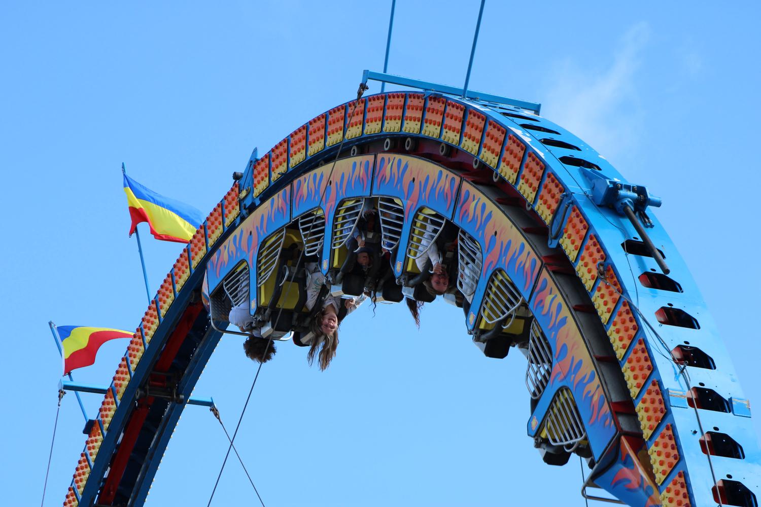 A brightly colored blue and yellow carnival ride with flame decals has riders hanging upside down against a clear blue sky. One person smiles directly at the camera while inverted.