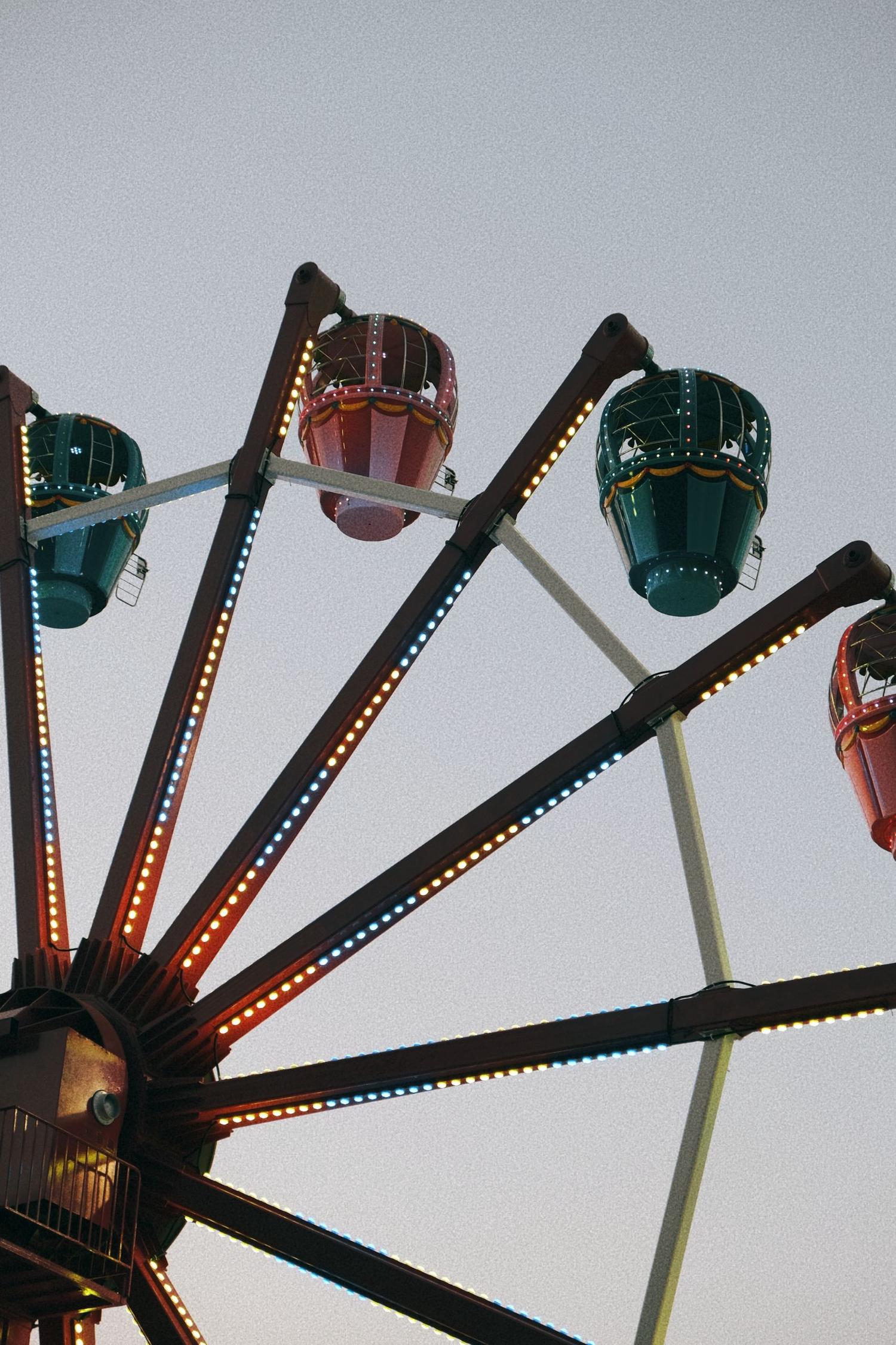 A section of a Ferris wheel against a grey sky, featuring red and teal passenger gondolas. Its dark spokes are illuminated with rows of alternating yellow and blue lights.