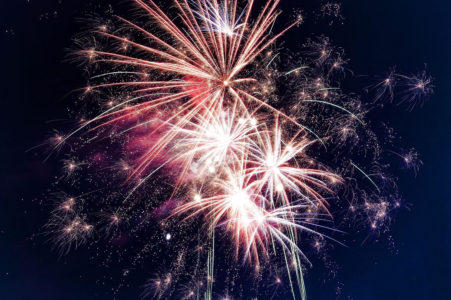 Multiple colorful fireworks burst across a dark night sky, with radiant streaks of red, white, and subtle green light creating a festive display.