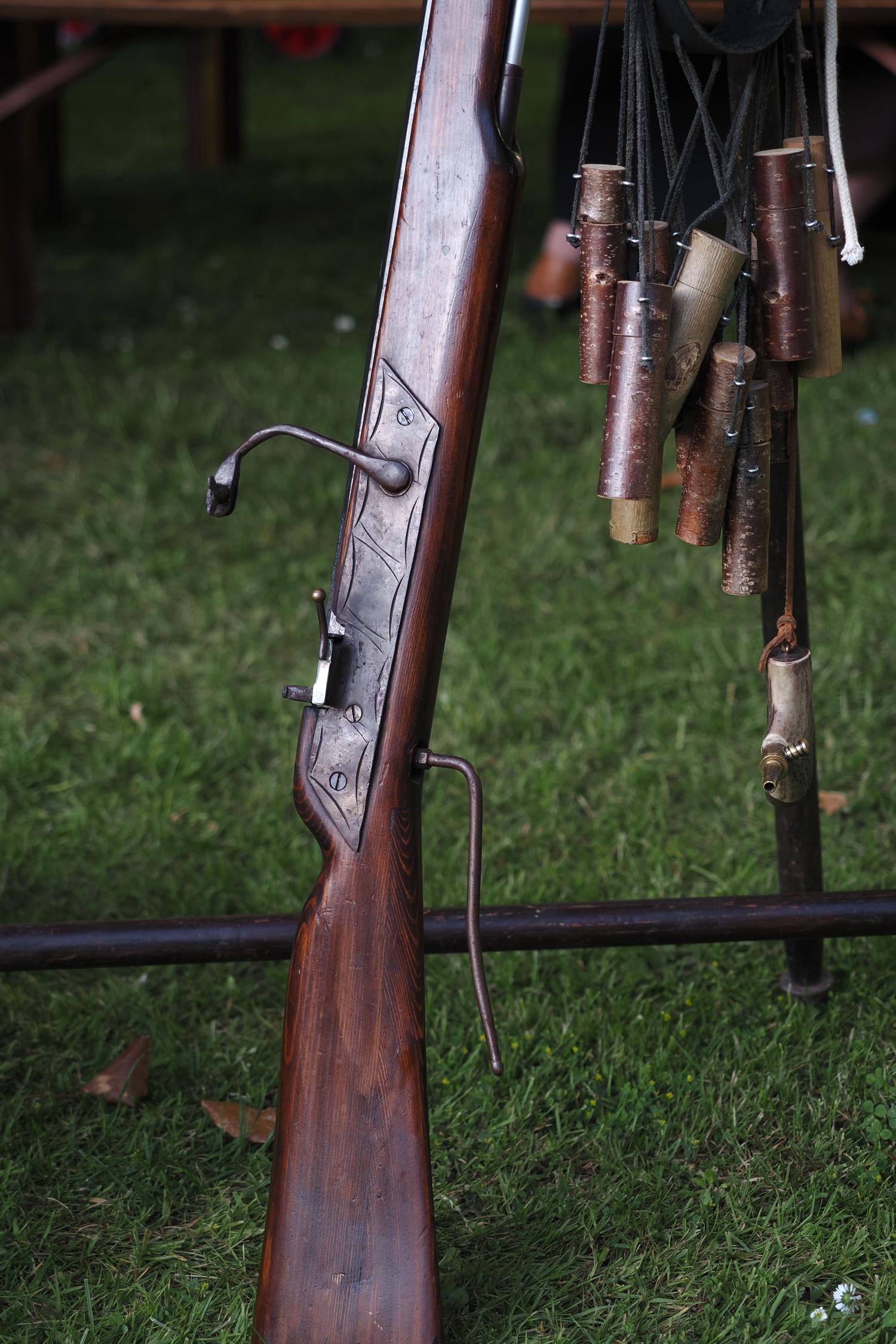 A dark wooden musket with a metal firing mechanism and long ramrod rests on green grass. Nearby hang several cylindrical wooden powder charges and a brass-tipped primer.