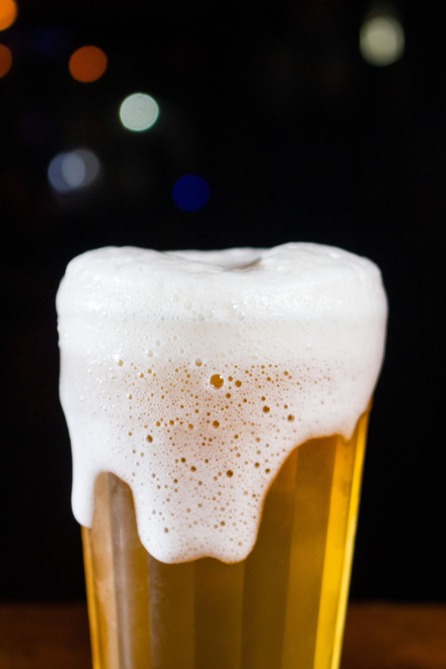A close-up of a golden amber beer in a glass with a generous, overflowing white foam head against a dark background with blurred colorful lights.