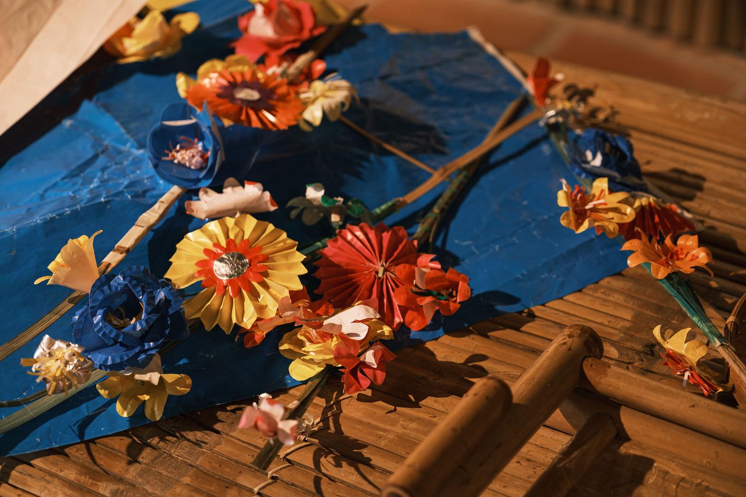 Colorful handmade paper flowers in red, yellow, blue, and orange are artfully arranged on a blue surface, which rests on a bamboo mat. The festive decorations include some flowers attached to slender sticks.