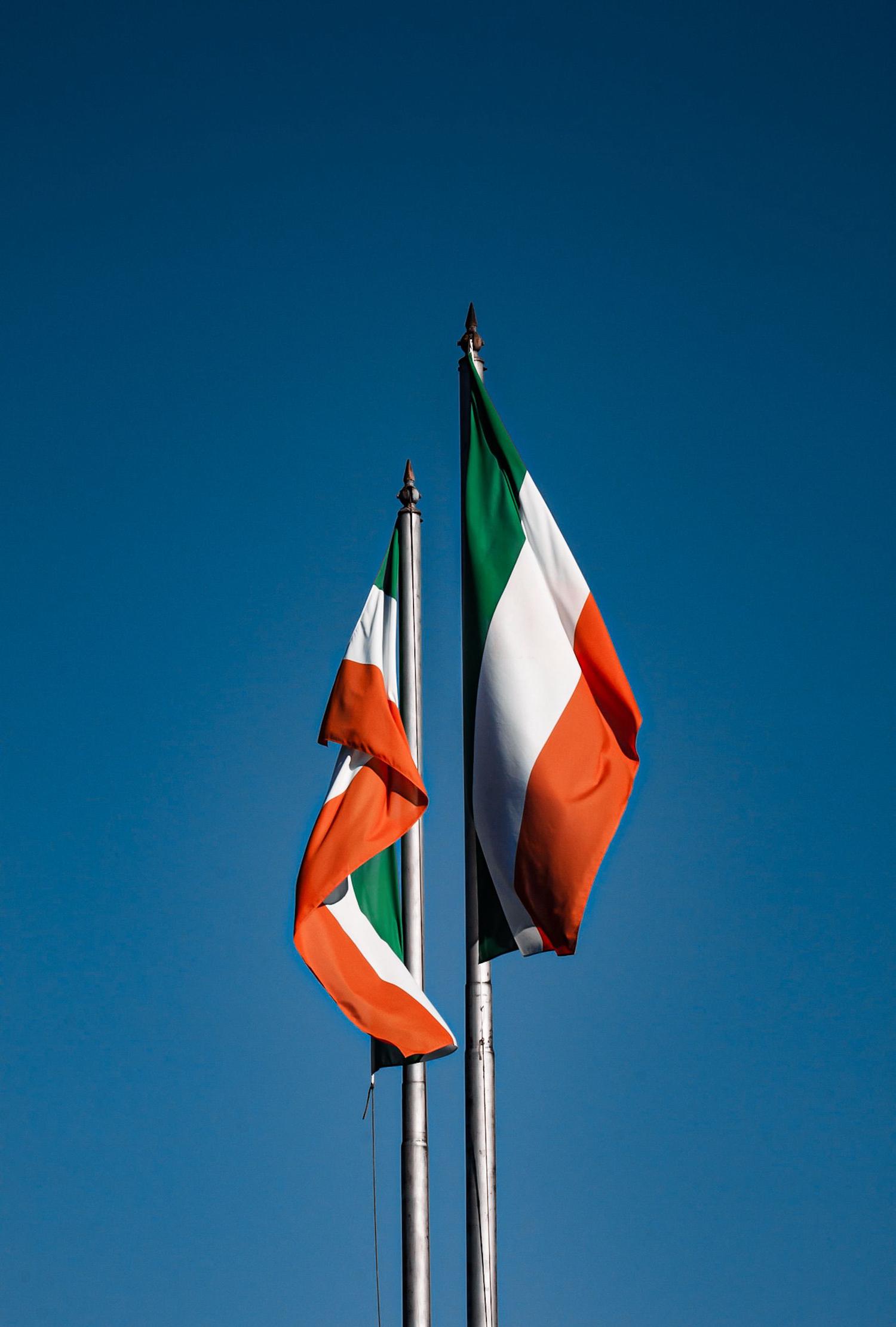 Two Irish flags, featuring vertical green, white, and orange stripes, fly on separate flagpoles against a bright blue sky. One flag is unfurled and blowing, while the other is partially wrapped around its pole.