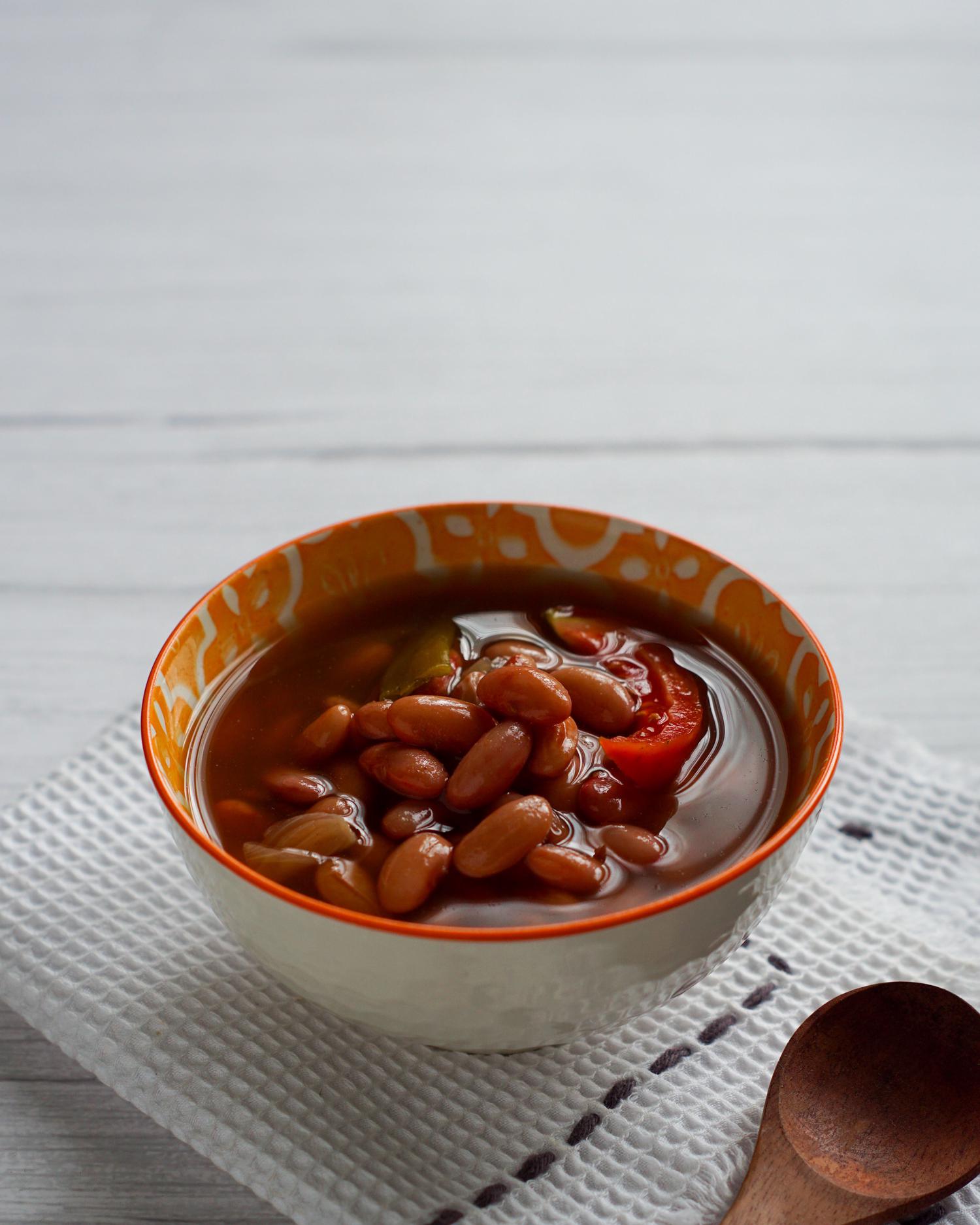 A bowl of reddish-brown bean soup with tomatoes and a rich broth sits on a white waffle-weave towel, with a wooden spoon visible nearby.