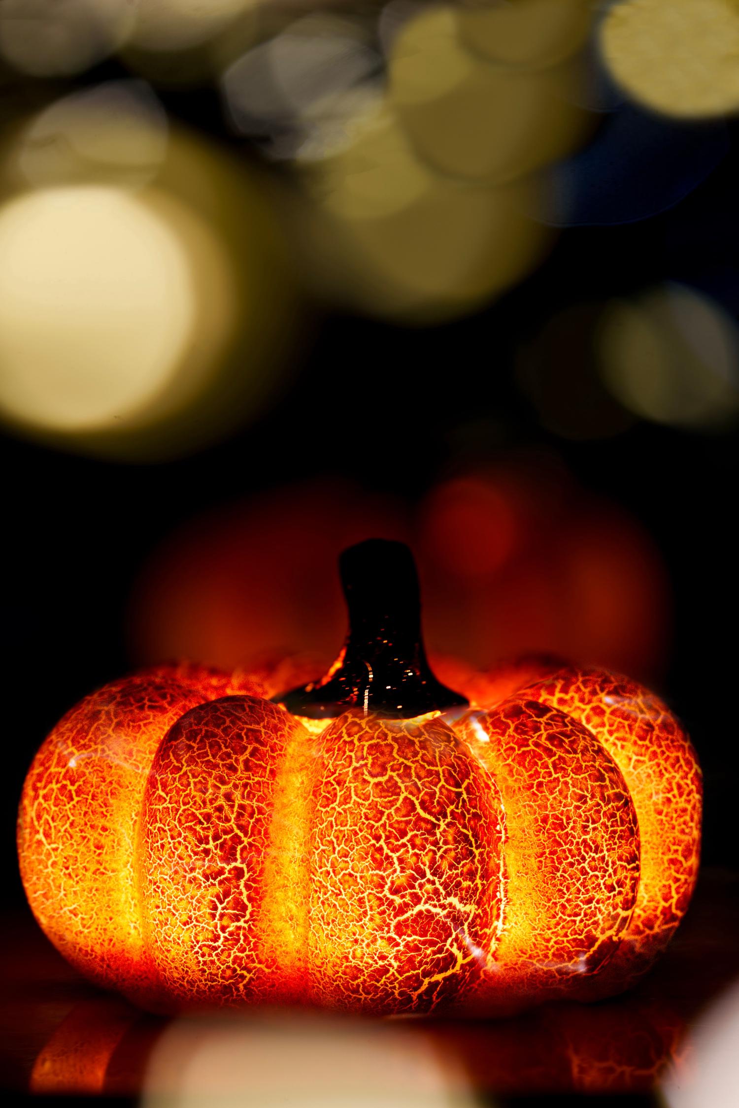 A decorative orange pumpkin glows brightly from within, showcasing a textured, crackled surface. It sits on a reflective surface against a dark, bokeh-filled background of warm, blurred lights.