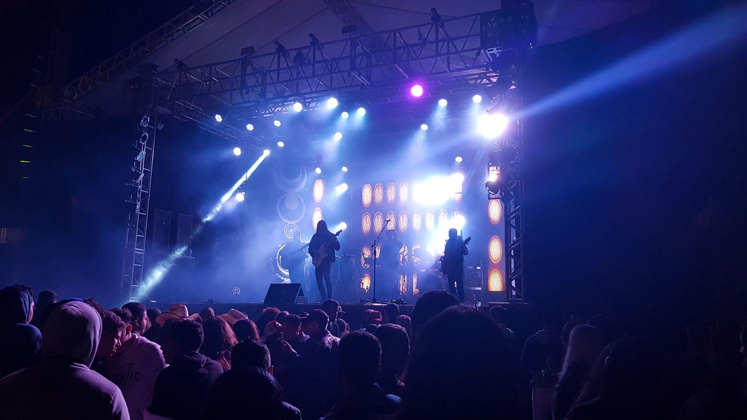 A band performs on a stage bathed in bright blue and purple lights, with a large, silhouetted audience watching from the foreground.