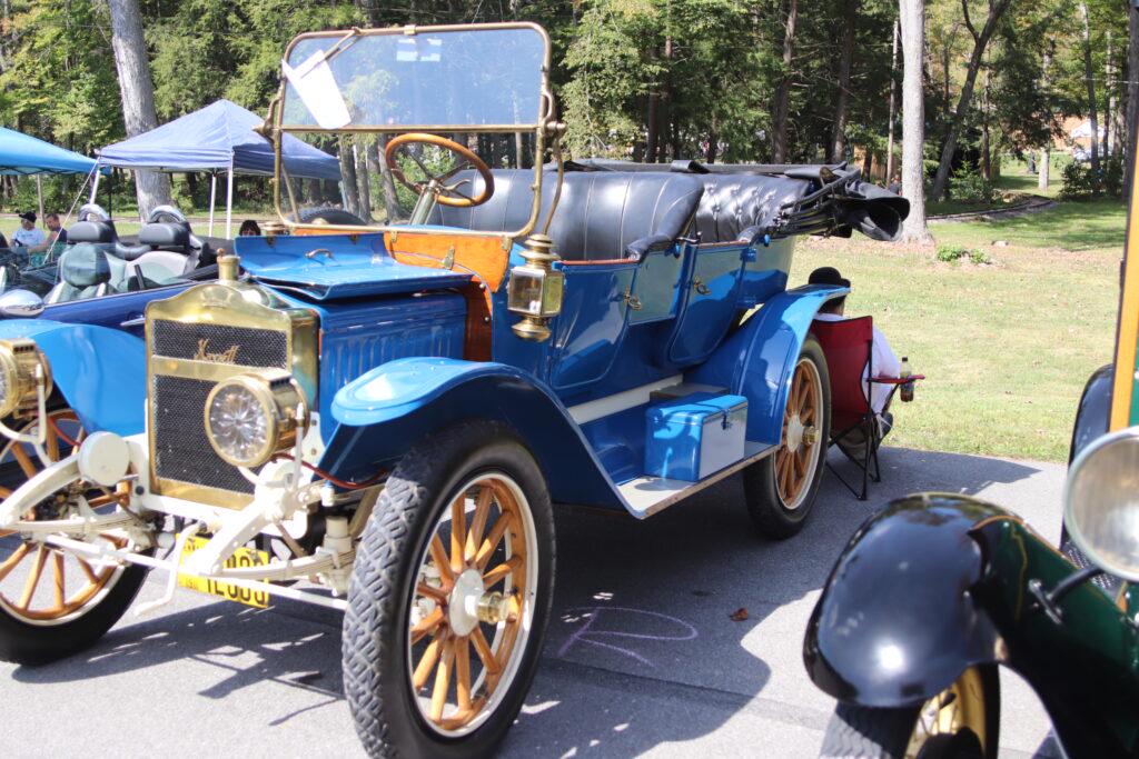 A vibrant blue vintage car with a gold grille, prominent headlights, and wooden spoke wheels is parked at an outdoor car show, with trees and tents in the background.