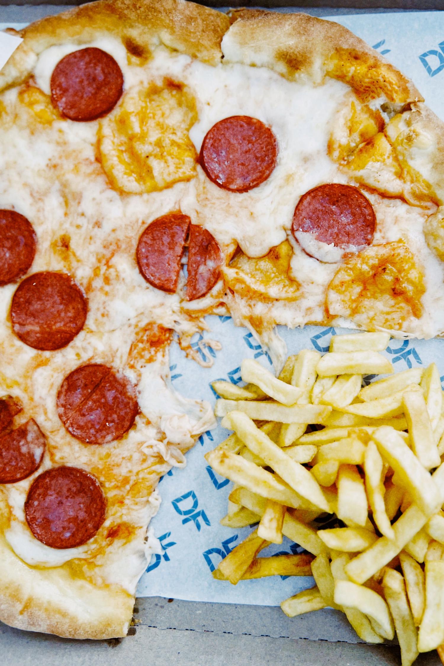 A partially eaten pepperoni and cheese pizza sits in a takeout box next to a mound of golden french fries. The box is lined with blue patterned paper.
