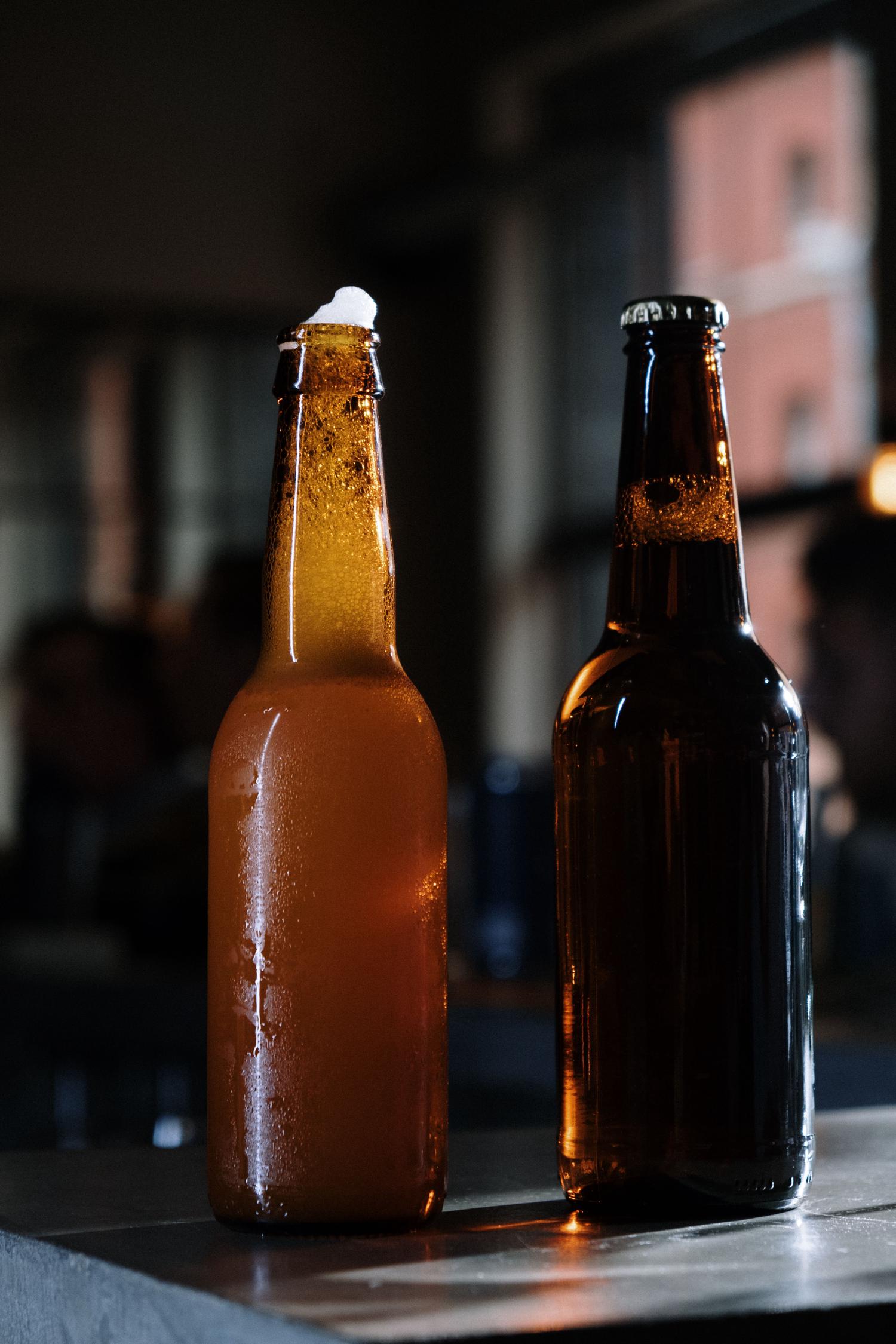Two beer bottles stand on a reflective surface in a dimly lit bar environment. The left bottle is a cold, amber beer with overflowing foam and condensation, next to a darker, capped bottle with visible bubbles.