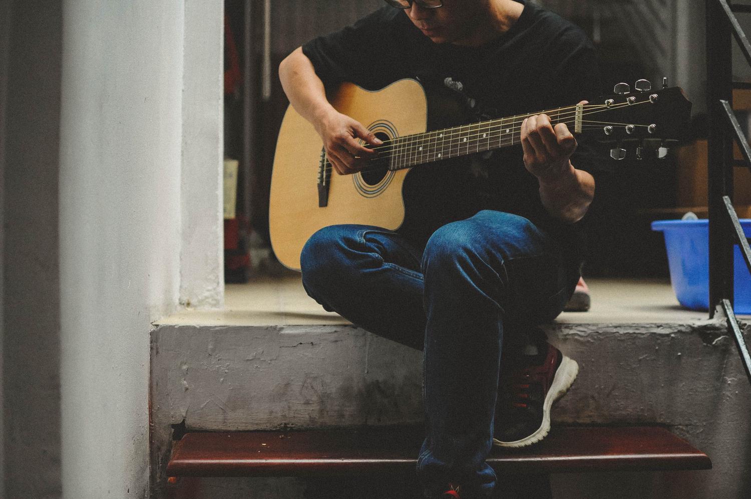 A person in a black shirt and blue jeans sits on steps, absorbed in playing an acoustic guitar.