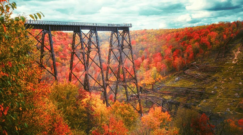 A tall, dark metal trestle bridge spans a deep valley vibrant with red, orange, and yellow autumn foliage. On the hillside below, remnants of a collapsed section of the bridge are scattered among the trees.