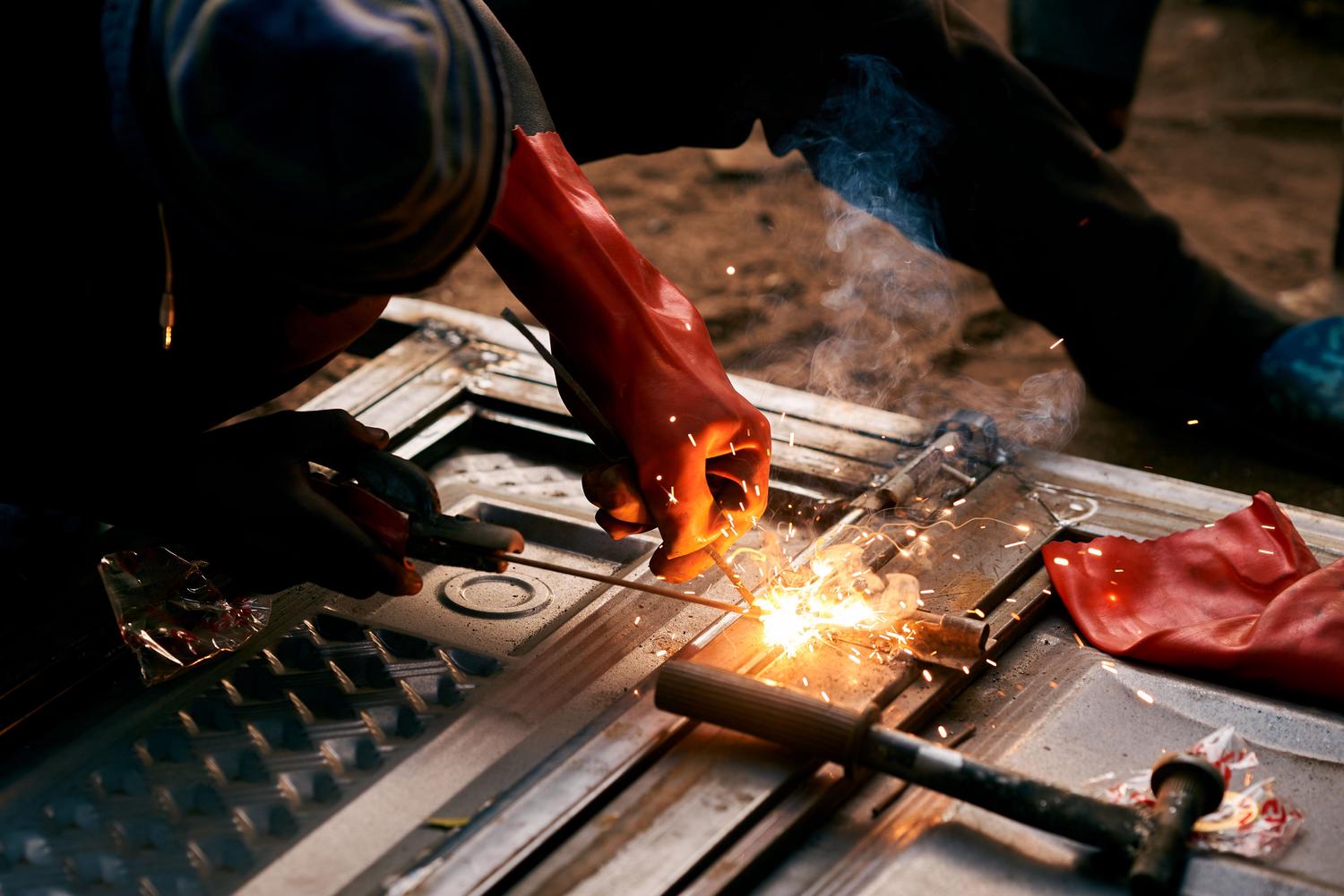 A person wearing a red glove uses welding equipment to join metal, creating bright sparks and a plume of smoke.