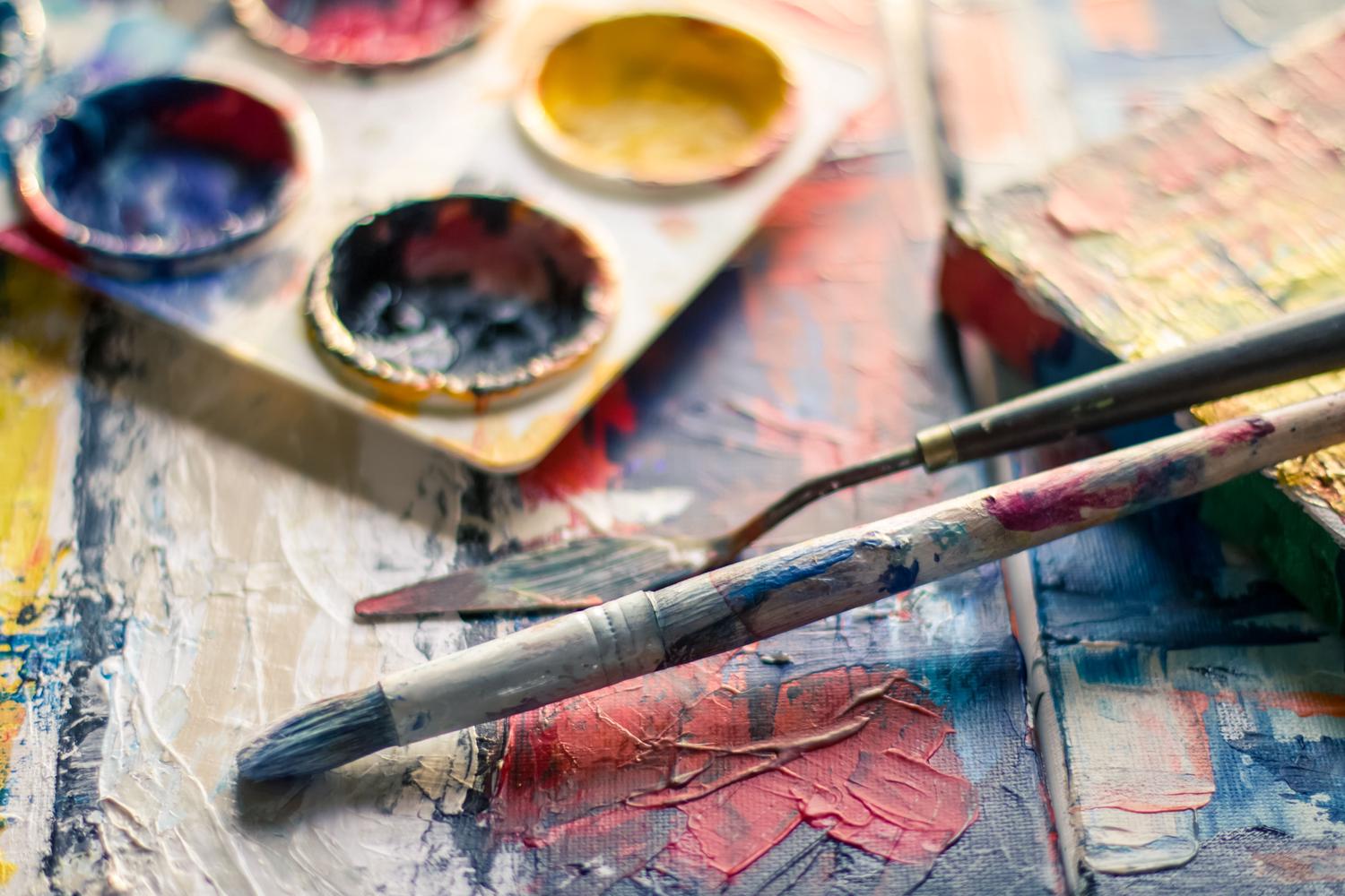 A paintbrush and a palette knife rest on a textured canvas covered in thick, vibrant strokes of red, blue, white, and yellow paint. An out-of-focus watercolor palette sits in the background.