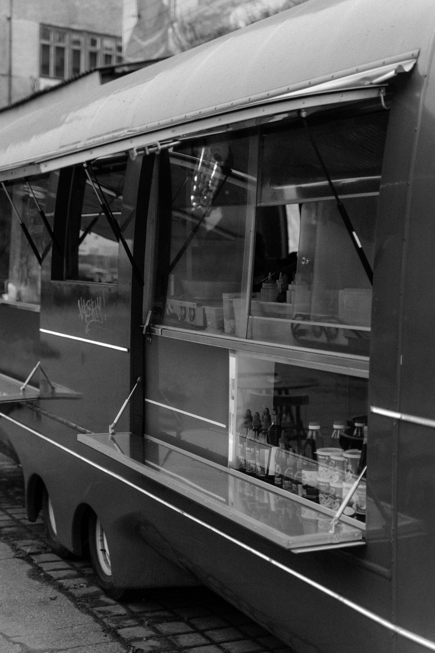 A black and white image showing the side of a food truck with its service windows open, revealing bottles and containers inside. The truck has a distinctive curved roof and a long, light-colored stripe along its lower body.