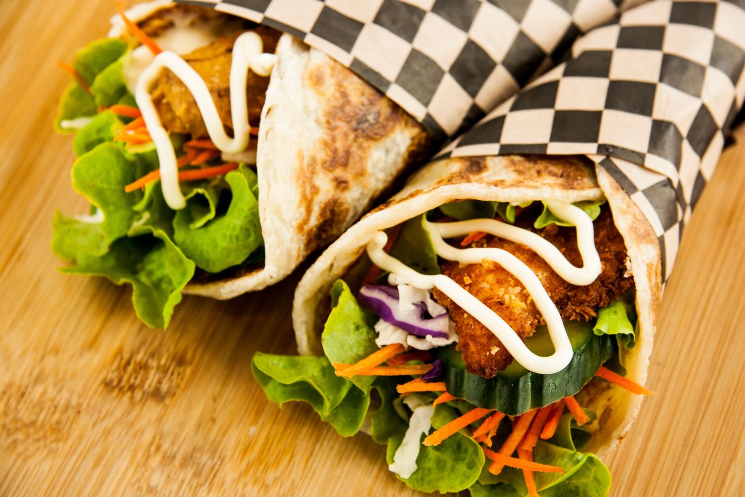 Two wraps filled with crispy golden protein, fresh green lettuce, shredded carrots, and white sauce are partially wrapped in black and white checkered paper and rest on a wooden surface.