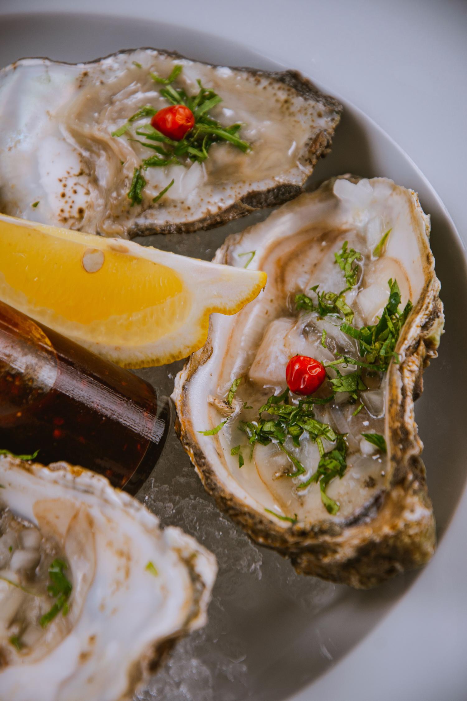 Fresh raw oysters on the half shell garnished with herbs, chopped onion, and red chilies, served on a bed of ice with a lemon wedge and a small bottle of sauce.
