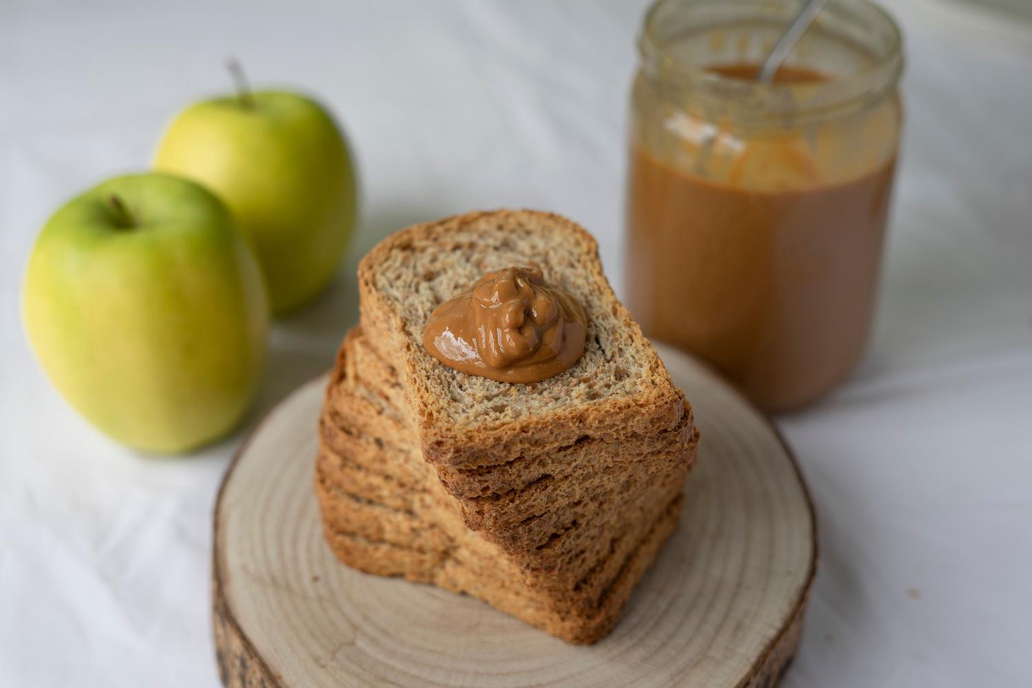 A stack of whole grain toast or rusks with a dollop of peanut butter on top sits on a wooden base, accompanied by two green apples and a jar of peanut butter in the background.