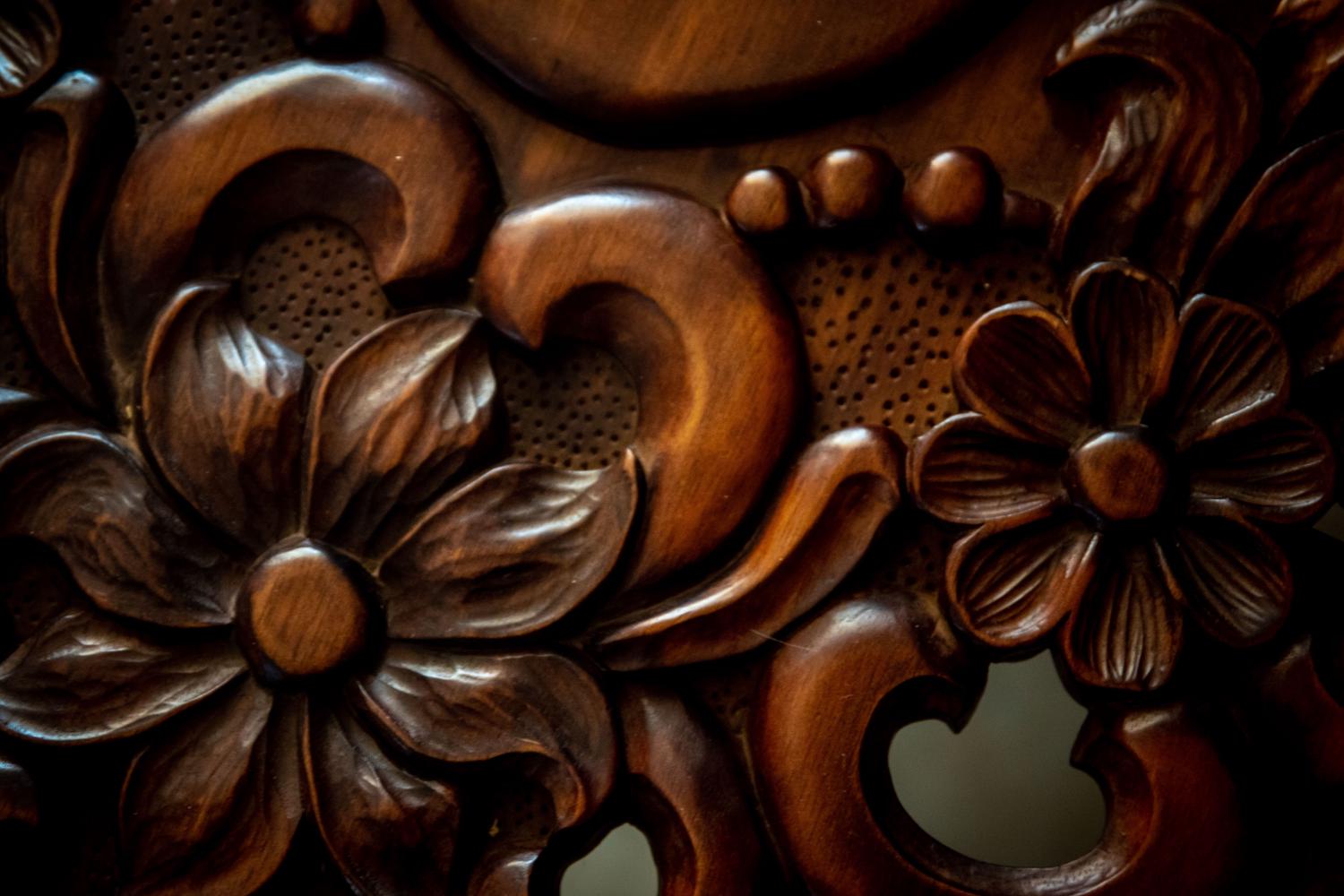 Close-up of intricate dark brown wooden carvings featuring ornate floral and leaf patterns. A stippled background texture contrasts with shiny, raised curves and deeply shadowed recesses.