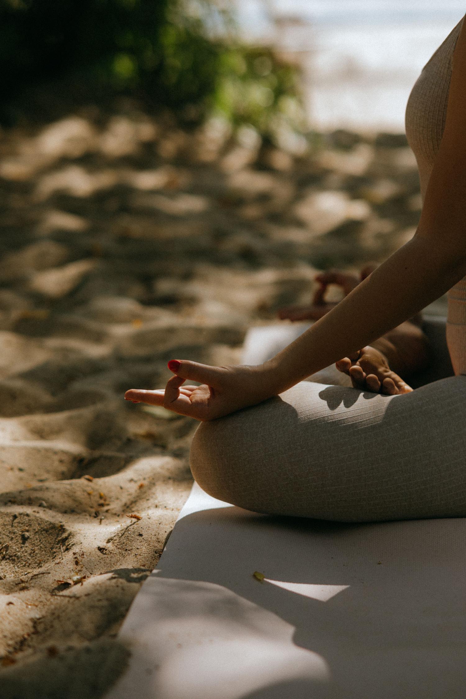 A person meditates outdoors in the lotus position on a yoga mat. Their hand, with red painted nails, rests on their knee in a mudra, illuminated by dappled sunlight on the sandy ground.