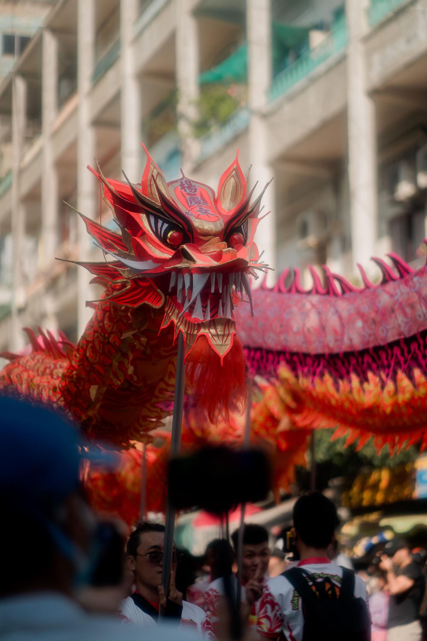 A vibrant red and gold dragon head with sharp teeth and large eyes is carried by people during a festive parade. Its detailed body, adorned with pink and gold patterns, extends behind, set against a blurred urban background.