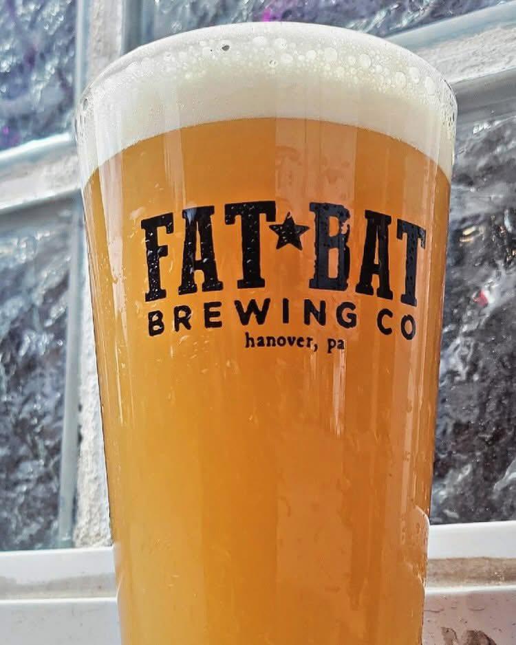 A glass of hazy, amber beer with a thick white head, featuring the black logo "FAT★BAT BREWING CO hanover, pa" on its side.