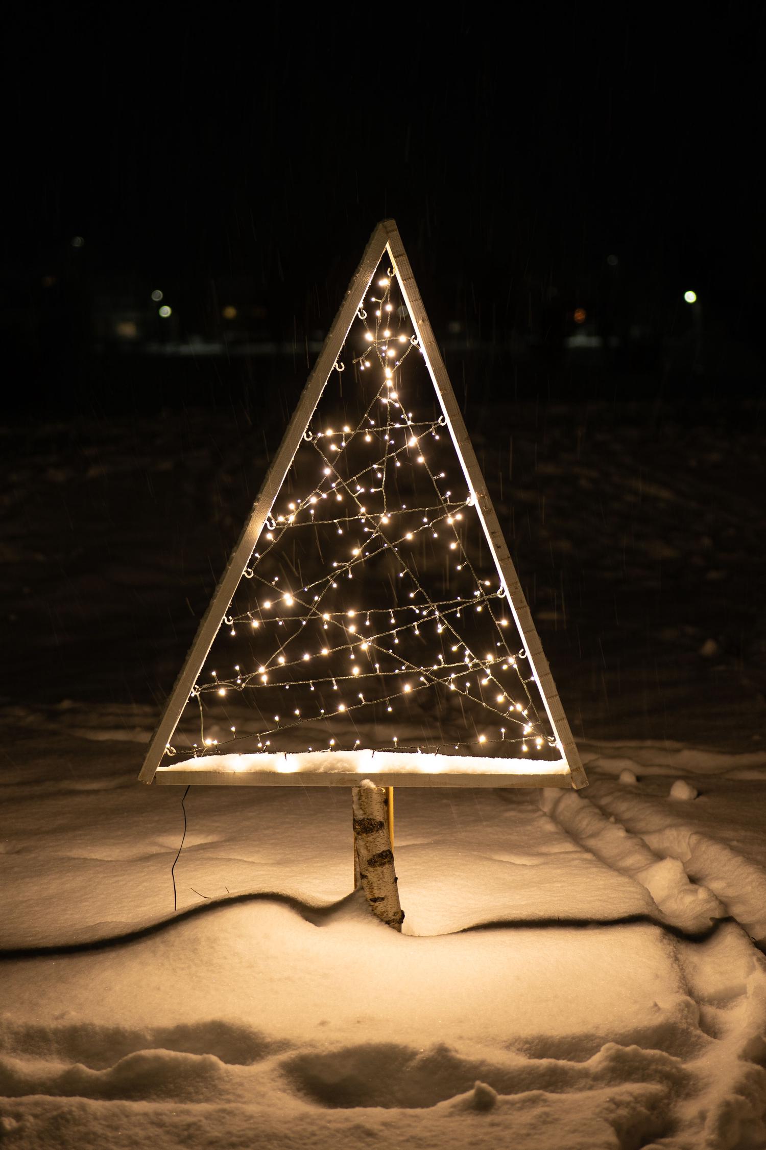 A modern, triangular Christmas tree made of a wooden frame with warm white string lights stands in fresh snow at night. A birch log acts as its trunk, casting a soft glow on the snowy ground.